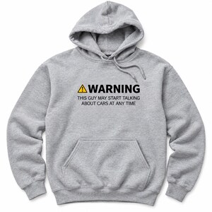 May include: A heather gray hooded sweatshirt featuring a front kangaroo pocket and a drawstring hood. The chest displays a yellow warning triangle icon followed by the black text: WARNING THIS GUY MAY START TALKING ABOUT CARS AT ANY TIME.