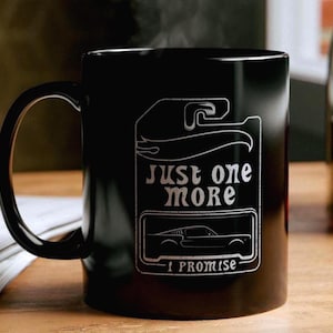 Op de afbeelding: Een zwarte keramische mok met een wit grafisch ontwerp dat lijkt op een verpakking van een speelgoedauto. Het ontwerp bevat de tekst "JUST ONE MORE" en "I PROMISE" met een lijntekening van het silhouet van een klassieke sportwagen.