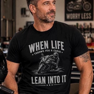 "Man in a garage wearing a black t-shirt printed with a sportbike racer graphic and the text 'When Life Throws You a Curve, Lean Into It.'"