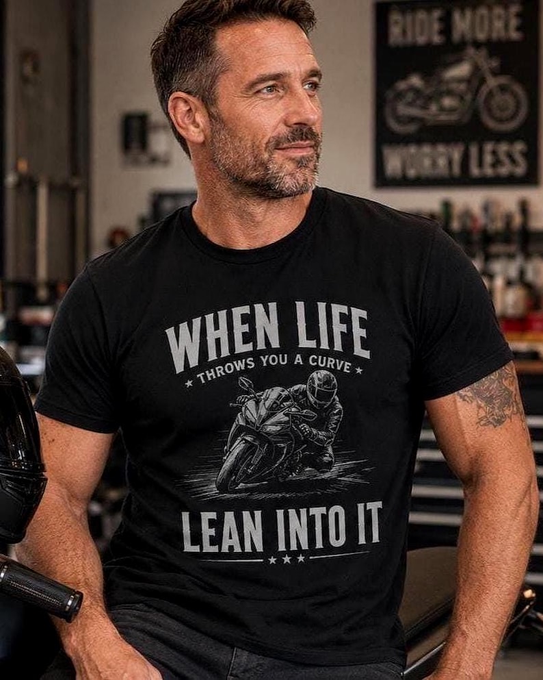 "Man in a garage wearing a black t-shirt printed with a sportbike racer graphic and the text 'When Life Throws You a Curve, Lean Into It.'"
