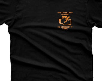 May include: A black short-sleeved t-shirt featuring an orange graphic on the left chest of an engine block with a lightning bolt symbol. The text reads: THIS LITTLE LIGHT OF MINE I'M GONNA LET IT SHINE. The inner neck label reads 365CAR MODS.