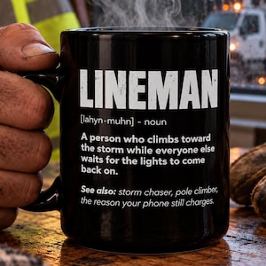 May include: A black ceramic mug featuring white text that defines the word LINEMAN as a person who climbs toward the storm while everyone else waits for the lights to come back on, with additional text including storm chaser and pole climber.