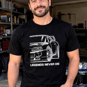May include: A black short-sleeve t-shirt featuring a white line art graphic of a sports car on the front. Below the car illustration, the text reads LEGENDS NEVER DIE in a bold, capitalized white font. The shirt has a crew neckline.