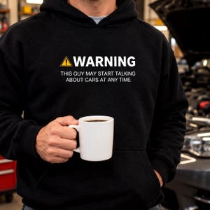 Close up of a black car guy hoodie with a warning design, worn in a home garage with a car behind him