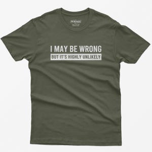 May include: An olive green short-sleeve t-shirt featuring the white text "I MAY BE WRONG BUT IT'S HIGHLY UNLIKELY" printed across the chest. The shirt has a crew neckline and a label inside the collar indicating 100% cotton and made in the USA.
