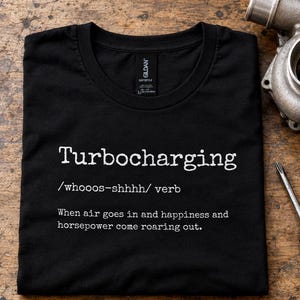 May include: A black t-shirt featuring white text that reads: Turbocharging /whooos-shhhh/ verb When air goes in and happiness and horsepower come roaring out. The shirt has a crew neckline and a Gildan brand label visible at the inner collar.