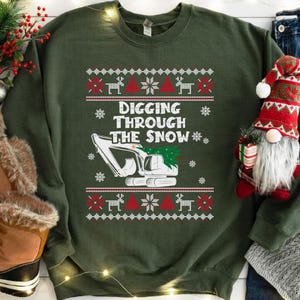 Peut inclure: Un sweat-shirt vert olive avec un motif festif. Le graphisme représente une excavatrice blanche avec un sapin de Noël, des flocons de neige et le texte "DIGGING THROUGH THE SNOW". Des motifs de rennes et d'arbres rouges et blancs complètent le thème des fêtes.