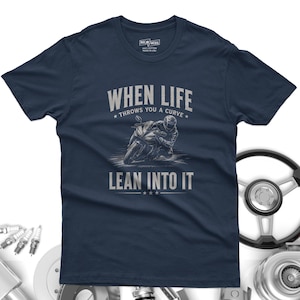 May include: A navy blue short-sleeve t-shirt featuring a graphic of a motorcyclist leaning into a curve. The text reads "WHEN LIFE THROWS YOU A CURVE LEAN INTO IT" in light gray lettering. The shirt has a crew neck and a printed interior neck label.