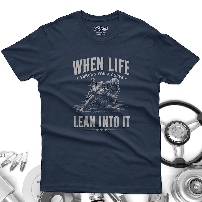 May include: A navy blue short-sleeve t-shirt featuring a graphic of a motorcyclist leaning into a curve. The text reads "WHEN LIFE THROWS YOU A CURVE LEAN INTO IT" in light gray lettering. The shirt has a crew neck and a printed interior neck label.