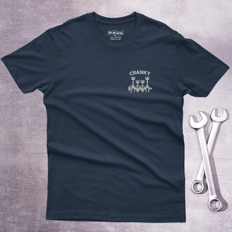 Op de afbeelding: Een marineblauw t-shirt met korte mouwen en een kleine witte afbeelding op de linkerborst. De afbeelding toont een krukas met zuigers en de tekst CRANKY erboven. Het shirt heeft een ronde hals en een label in de kraag met CAR MODS.