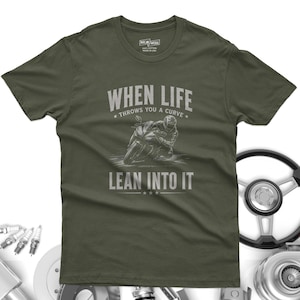 May include: An olive green short-sleeve t-shirt featuring a graphic of a motorcyclist leaning into a curve. The text reads "WHEN LIFE THROWS YOU A CURVE LEAN INTO IT" in a distressed white font. The shirt has a crew neck and a standard fit.