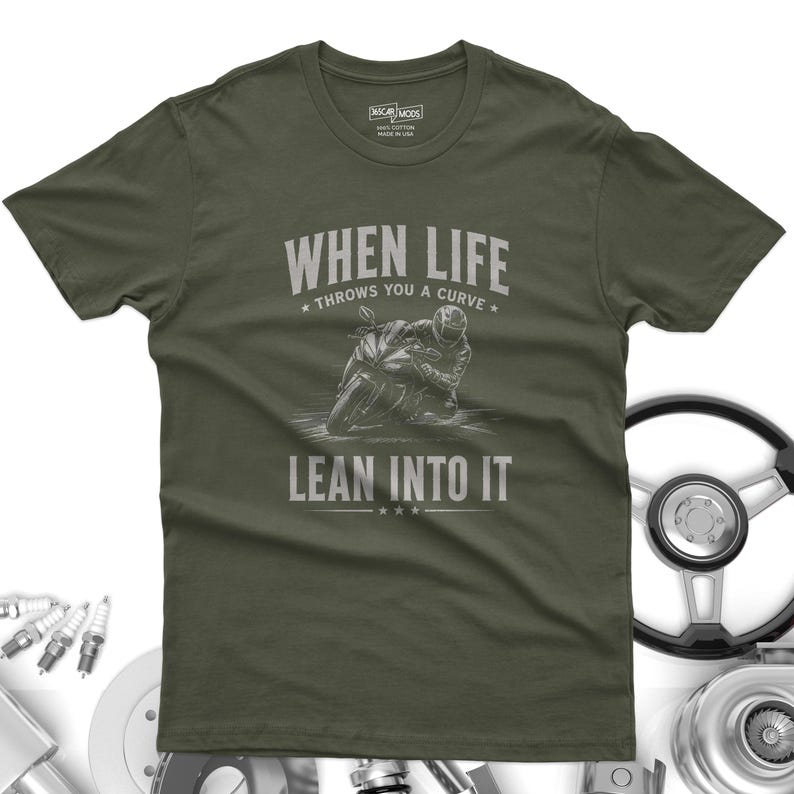 May include: An olive green short-sleeve t-shirt featuring a graphic of a motorcyclist leaning into a curve. The text reads "WHEN LIFE THROWS YOU A CURVE LEAN INTO IT" in a distressed white font. The shirt has a crew neck and a standard fit.
