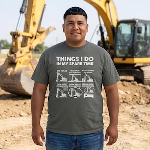 May include: A man wearing a gray t-shirt with the text "THINGS I DO IN MY SPARE TIME" and excavator-themed graphics. The background features a yellow excavator and a construction site.