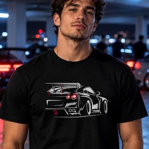 Puede incluir: Una camiseta negra de manga corta con una ilustración gráfica en blanco de la parte trasera de un coche deportivo. El diseño del coche incluye un alerón trasero prominente, guardabarros anchos, luces traseras rojas y un detalle rojo.