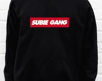 May include: A black hooded sweatshirt featuring a rectangular red graphic on the chest with the white text SUBIE GANG printed in a bold, sans-serif font. The garment includes a front kangaroo pocket, long sleeves, and a drawstring hood.