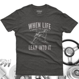 May include: A charcoal gray short-sleeve t-shirt featuring a graphic of a motorcyclist leaning into a curve. The text reads "WHEN LIFE THROWS YOU A CURVE LEAN INTO IT" in a light gray font. The shirt has a crew neck and a standard fit design.