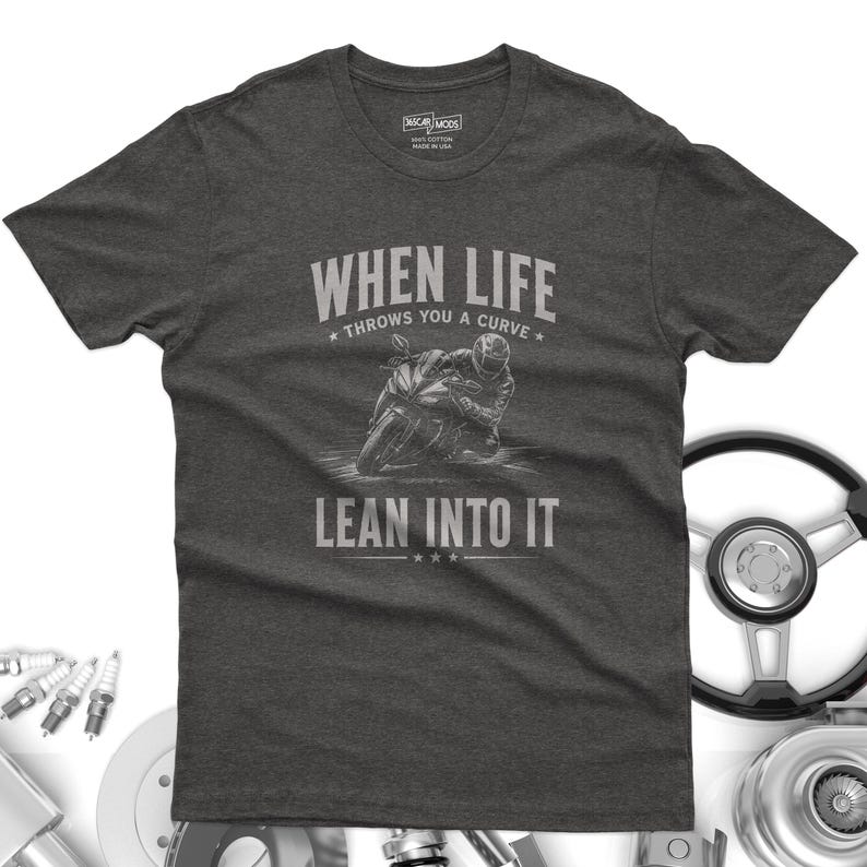 May include: A charcoal gray short-sleeve t-shirt featuring a graphic of a motorcyclist leaning into a curve. The text reads "WHEN LIFE THROWS YOU A CURVE LEAN INTO IT" in a light gray font. The shirt has a crew neck and a standard fit design.