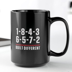 Op de afbeelding: Een zwarte keramische mok met witte tekst die 1-8-4-3 6-5-7-2 leest, een dunne rode horizontale lijn eronder en de woorden BUILT DIFFERENT in witte hoofdletters onder de lijn gedrukt. De mok heeft een groot handvat aan de rechterkant.