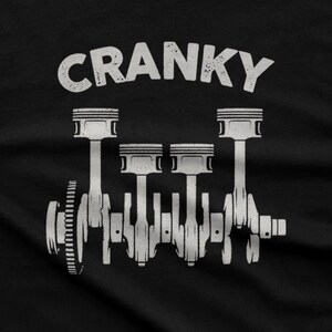 Op de afbeelding: Een zwart t-shirt met een witte grafische print van een viercilinder motor-krukas met zuigers. Het woord CRANKY is in een verweerd wit lettertype boven de mechanische illustratie op de stof gecentreerd afgedrukt.