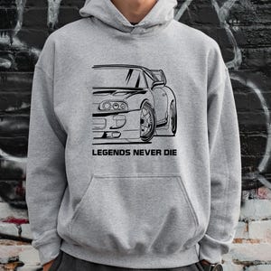 Puede incluir: Una sudadera con capucha gris con un dibujo lineal en blanco y negro de un coche deportivo. El texto "LEGENDS NEVER DIE" est&aacute; impreso debajo del gr&aacute;fico del coche. La sudadera tiene un bolsillo delantero y una capucha.