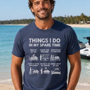 以下が含まれることがあります： 白のテキストとグラフィックがプリントされた、ヘザーネイビーブルーの半袖Tシャツ。デザインには「THINGS I DO IN MY SPARE TIME」という文字と、6つのアイコンおよびキャプション「RIDE MY JET SKI」「CHECK THE WEATHER」「WAIT IN LINE AT THE RAMP」「FLUSH IT AFTER EVERY RIDE」「WATCH IT SIT ALL WINTER」「DREAM ABOUT JET SKIS」が描かれています。