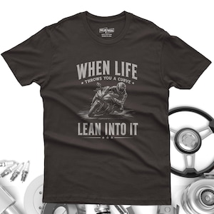 May include: A dark charcoal gray short-sleeve t-shirt featuring a graphic of a motorcyclist leaning into a turn. The text reads "WHEN LIFE THROWS YOU A CURVE LEAN INTO IT" in a light gray font. The shirt has a crew neck and a standard fit.