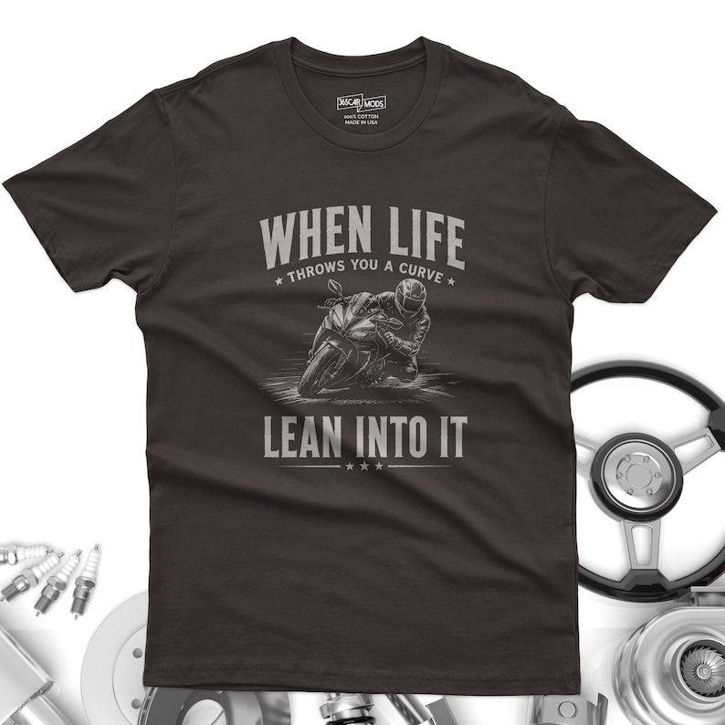 May include: A dark charcoal gray short-sleeve t-shirt featuring a graphic of a motorcyclist leaning into a turn. The text reads "WHEN LIFE THROWS YOU A CURVE LEAN INTO IT" in a light gray font. The shirt has a crew neck and a standard fit.