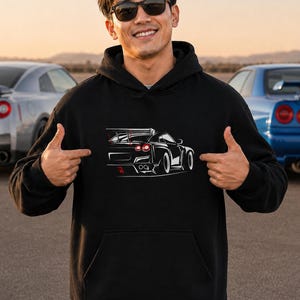 Puede incluir: Una sudadera negra con capucha que presenta un gráfico de líneas blancas de la vista trasera de un coche deportivo. El diseño incluye un alerón trasero prominente, luces traseras circulares y un difusor, con pequeños detalles en color rojo.