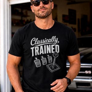 以下が含まれることがあります： 黒の半袖Tシャツ。白い文字で「Classically TRAINED」と書かれ、その下に3つの車のペダルと、6速シフトパターンが描かれたマニュアルトランスミッションのシフトレバーのグラフィックイラストがプリントされています。