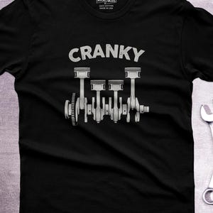 Könnte beinhalten: Ein schwarzes Kurzarm-T-Shirt mit einer grauen Grafik einer Kurbelwelle mit vier Kolben. Das Wort CRANKY ist in Grau über den Motorkomponenten aufgedruckt. Das Innenetikett zeigt den Text 365CAR MODS 100% COTTON MADE IN USA.