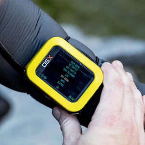 Peut inclure: Un ordinateur de plongée jaune et noir monté au poignet avec un écran numérique affichant les options de menu : Menu principal, Commutateur SP, Commutateur de gaz, Planificateur de plongée, Journal, Menu de configuration, Informations, Ardoises. L'ordinateur possède un bouton circulaire sur le côté gauche et un bouton rectangulaire sur le côté droit.