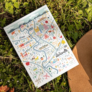 Pu&ograve; includere: Una mappa illustrata colorata di Asheville, Carolina del Nord, con il testo "Asheville, Lord of the Sky" e "The Crafted Company". La mappa presenta monumenti, quartieri e punti di interesse.