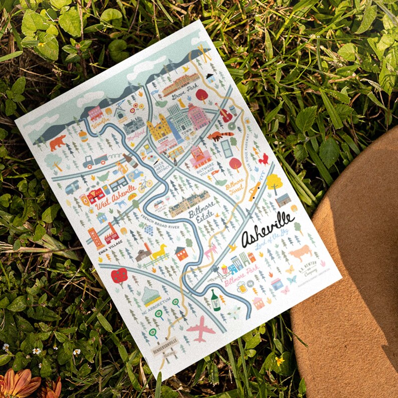 Pu&ograve; includere: Una mappa illustrata colorata di Asheville, Carolina del Nord, con il testo "Asheville, Lord of the Sky" e "The Crafted Company". La mappa presenta monumenti, quartieri e punti di interesse.