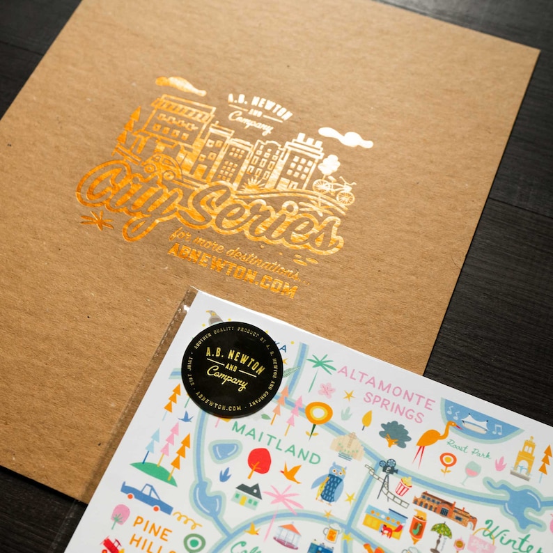 May include: A brown paper envelope with a gold foil design of a city skyline and the text "A.B. Newton Company City Series for more destinations ABNEWTON.COM". The envelope is partially open and a colorful illustrated map of a city is visible inside.