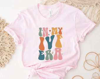 Puede incluir: Camiseta rosa con un gráfico de estilo retro que dice "In My Era" en letras de colores con corazones.