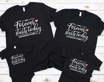May include: A set of four black t-shirts with the text "Our Forever Starts Today" and a pink heart. The shirts are in different sizes for adults and children. The text "Love Makes a Family" is also included on the shirts.