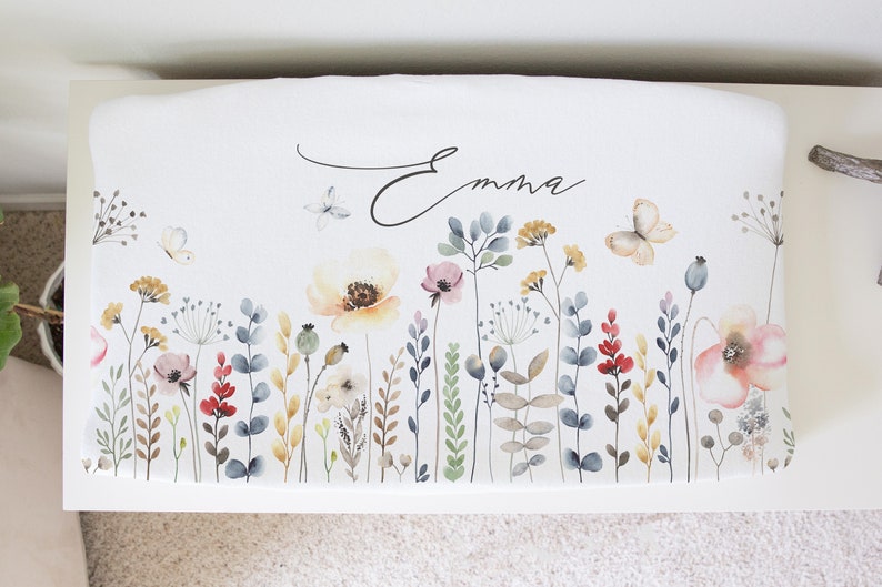 May include: White bench cushion with a floral design featuring butterflies, flowers, and greenery in shades of pink, yellow, red, and blue. The name "Emma" is written in cursive on the cushion.