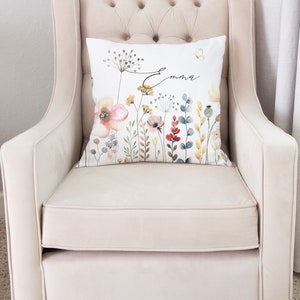 Puede incluir: Una almohada blanca con un dise&ntilde;o floral de acuarela y el nombre "Emma" en escritura cursiva negra. La almohada est&aacute; sentada en un sill&oacute;n beige claro.