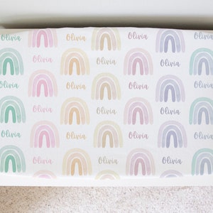 May include: A white crib sheet with a repeating pattern of pastel rainbows and the name "Olivia" printed on each rainbow.