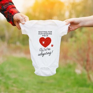 May include: White baby onesie with a red heart puzzle piece design. The text reads "Waiting for our missing piece. We're adopting!"