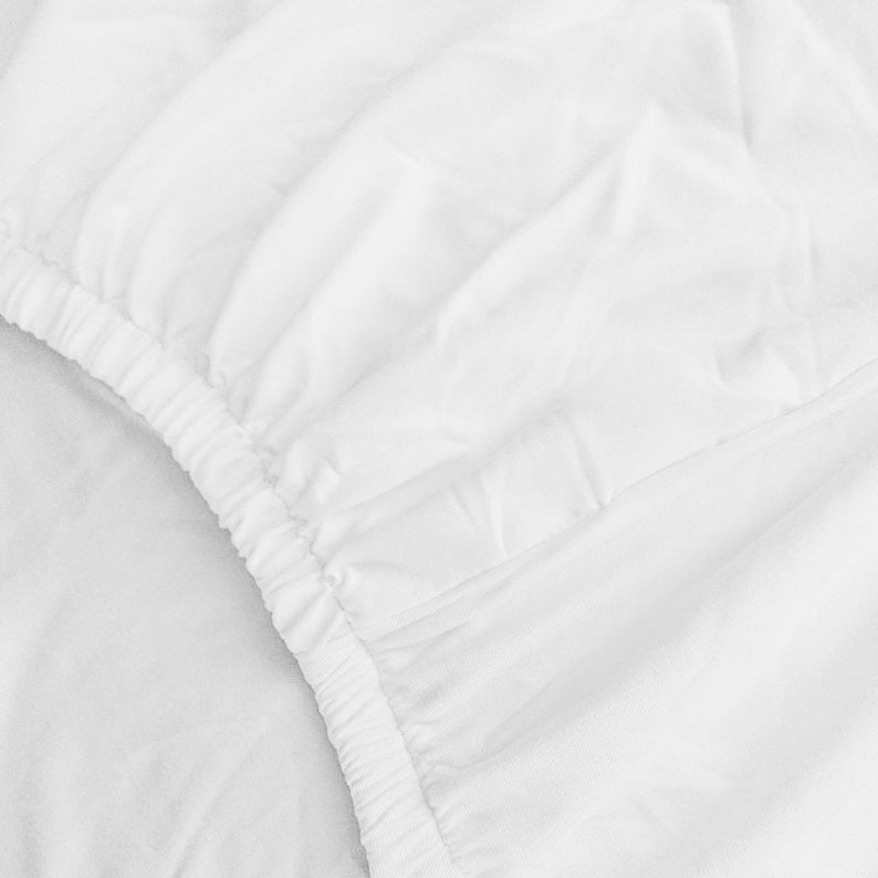 Puede incluir: Primer plano de una s&aacute;bana bajera blanca con borde el&aacute;stico. La s&aacute;bana est&aacute; hecha de una tela suave y lisa.