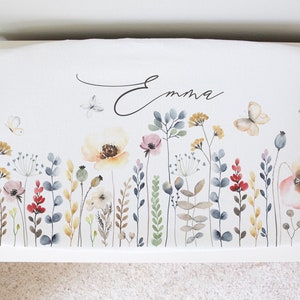May include: White bench cushion with a floral design featuring butterflies, flowers, and greenery in shades of pink, yellow, red, and blue. The name "Emma" is written in cursive on the cushion.