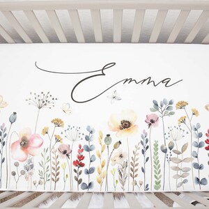 May include: White crib sheet with a watercolor floral design featuring butterflies and the name "Emma" in cursive script.