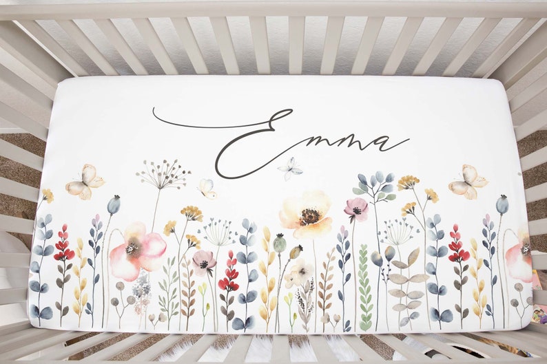 May include: White crib sheet with a watercolor floral design featuring butterflies and the name "Emma" in cursive script.