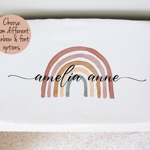 May include: A white crib sheet with a watercolor rainbow in shades of brown, pink, and beige. The name "amelia anne" is written in cursive script below the rainbow.