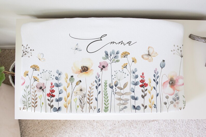 Puede incluir: Coj&iacute;n de banco blanco con un dise&ntilde;o floral que presenta mariposas, flores y vegetaci&oacute;n en tonos de rosa, amarillo, rojo y azul. El nombre "Emma" est&aacute; escrito en cursiva en el coj&iacute;n.