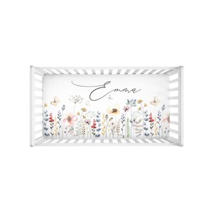 Puede incluir: S&aacute;bana de cuna blanca con un dise&ntilde;o floral de acuarela con mariposas y el nombre "Emma" en letra cursiva.