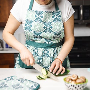 May include: A person wearing a white t-shirt and a teal and green patterned apron, slicing zucchini on a white countertop. A bowl of mushrooms and a bowl of vegetables are nearby. A matching pot holder is on the counter.