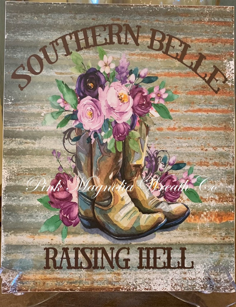 May include: A rustic wooden sign with the text "Southern Belle Raising Hell" in brown lettering. The sign features a watercolor painting of two brown cowboy boots with a bouquet of pink, purple, and white flowers.