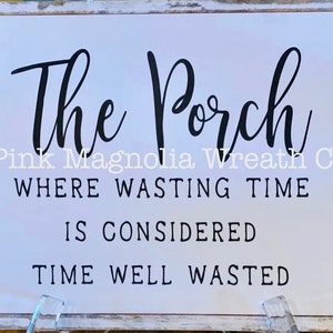 Pu&ograve; includere: Un cartello di legno bianco con testo nero che recita "The Porch Pink Magnolia Wreath Co. Where wasting time is considered time well wasted."